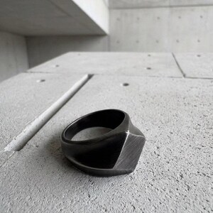 May include: A matte black ring with a geometric, angular design featuring a prominent, flat-faced square front section. The band has a smooth, brushed texture and a solid, thick construction that tapers slightly toward the back of the ring.