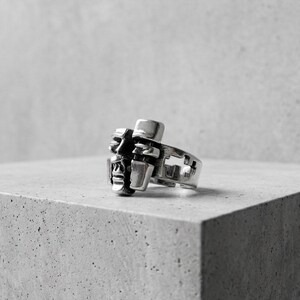 May include: A silver-toned metal ring featuring a sculptural design of two stylized faces positioned side-by-side. The band of the ring includes a series of rectangular cutouts along the side, and the metal has a polished, reflective finish.