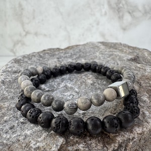 May include: Two beaded bracelets. The top bracelet features gray and white stone beads with a silver square accent. The bottom bracelet has black lava stone beads. Both bracelets are resting on a gray stone surface.