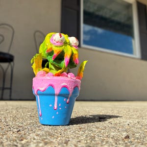 May include: A hand-painted sculpture of a green, multi-eyed creature emerging from a blue flower pot. The pot features a pink, dripping paint effect around the rim. The creature has bright green skin, yellow accents, and two prominent white eyeballs.