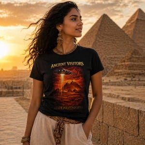 Może przedstawiać: Czarny t-shirt z krótkim rękawem z nadrukiem graficznym przedstawiającym latający spodek unoszący się nad piramidami z wiązką światła. Napis "ANCIENT VISITORS" jest nadrukowany złotym kolorem nad grafiką, a na dole znajdują się ozdobne symbole.