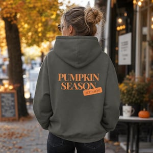 Könnte beinhalten: Ein dunkelgrauer Kapuzenpullover mit dem orangefarbenen Schriftzug PUMPKIN SEASON ACTIVATED auf der Rückseite. Der Hoodie hat lange Ärmel, eine große Kapuze und eine Kängurutasche, wobei der Text in einer fetten Schriftart dargestellt ist.