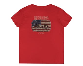 May include: A red short-sleeved t-shirt featuring a graphic on the back. The graphic displays a distressed American flag with the text "THE STARS CHANGED. THE PROMISE DIDN'T." above it, the word "Liberty." in the centre, and "1776-2026" below.