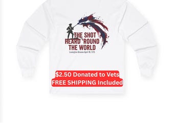 Puede incluir: Una camiseta blanca de manga larga con un gráfico en la parte posterior. El gráfico muestra la silueta de un soldado con un mosquete, acompañado de salpicaduras de pintura roja y azul y el texto: THE SHOT HEARD ROUND THE WORLD Lexington Greene April 19, 1775.