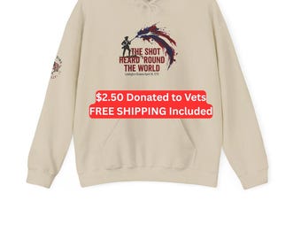 Puede incluir: Una sudadera con capucha de color beige que presenta un gráfico de un soldado y un efecto de salpicadura de pintura roja, blanca y azul. El texto dice: THE SHOT HEARD 'ROUND THE WORLD Lexington Green April 19, 1775. Un pequeño emblema es visible en la manga derecha.