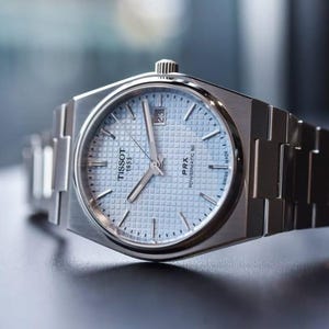 May include: A silver-toned stainless steel wristwatch featuring a light blue waffle-patterned dial, silver baton indices, and silver hands. The watch face displays the Tissot 1853 logo, PRX Powermatic 80 text, a date window at the three o'clock position.
