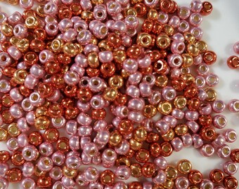 May include: A collection of small, round beads in shades of pink, gold, and copper. The beads are scattered on a white surface, creating a textured and colorful display. These beads are ideal for jewelry making and crafting projects.