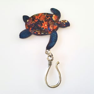 May include: A colorful, patterned sea turtle charm with a silver hook. The turtle is blue, black, and orange with a floral design.