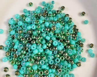 May include: A pile of turquoise and green glass seed beads. The beads are small and round, and they have a shiny finish.