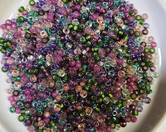 May include: A close-up shot of a white bowl filled with a variety of small, colorful beads. The beads are a mix of purple, green, pink, and iridescent colors. The beads are small and round, perfect for jewelry making or other crafts.