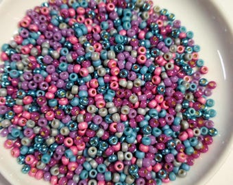 May include: A bowl of seed beads in shades of pink, purple, blue, and silver. The beads are small and round, and they are perfect for jewelry making or other craft projects.
