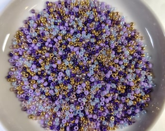 May include: A collection of small, colorful beads in a white bowl. The beads are a mix of purple, gold, and light blue. The beads are small and round, and they appear to be made of glass or plastic. The bowl is round and has a slightly raised edge.