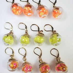 May include: Nine pairs of dangle earrings with glass spheres containing colorful fruit slices. The earrings are gold-toned and have a hook closure. The fruit slices are orange, yellow, and pink.