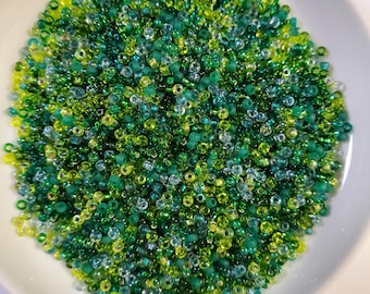 May include: A white ceramic bowl filled with a variety of small, colorful beads. The beads are primarily shades of green and yellow, creating a vibrant and textured appearance. The beads are likely used for crafting or jewelry making.