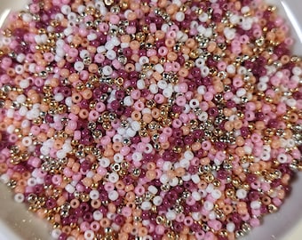 May include: A close-up shot of a collection of small, colorful beads in a ceramic dish. The beads are a mix of pink, white, peach, silver, and gold. The beads are scattered, creating a textured surface.