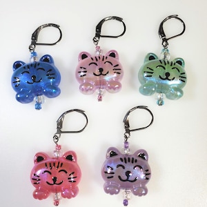 May include: Five colorful cat charm keychains with iridescent finishes. Each charm features a black outline of a cat face with a smiling expression. The charms are attached to silver key rings.