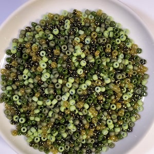May include: A selection of small, round beads in shades of green and gold, presented in a white dish. The beads vary in colour from light green to dark olive and metallic gold, creating a textured, natural look. Suitable for jewellery making and crafts.