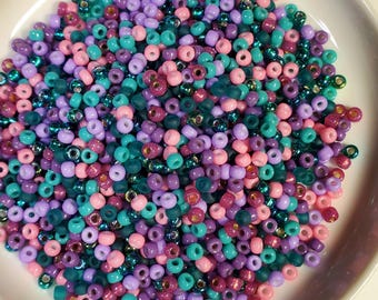 May include: A bowl filled with small, colorful glass beads in shades of pink, purple, teal, and green. The beads have a metallic sheen and are perfect for jewelry making or crafting.