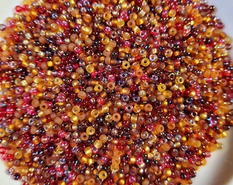 May include: A close-up shot of a pile of small, colorful beads in shades of red, gold, and brown. The beads are scattered in a white bowl, creating a textured and vibrant display. These beads are ideal for jewelry making and crafting.