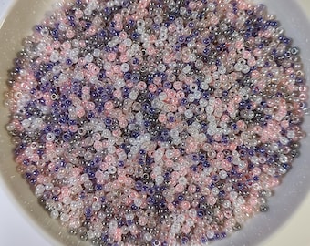 May include: A ceramic plate filled with a mix of small, round beads. The beads are in shades of pink, purple, white, and gray. The beads are scattered across the plate, creating a colorful and textured surface.
