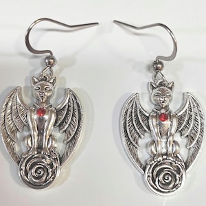 Puede incluir: Un par de pendientes plateados con un diseño de gato alado. Cada pendiente tiene una gema roja en el pecho y una rosa en la base. Los pendientes tienen cierre de gancho.