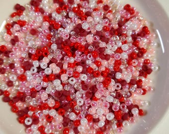 May include: A white bowl filled with a variety of small, round seed beads in shades of red, pink, and white.