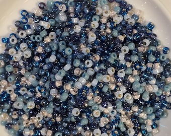 May include: A close-up shot of a white bowl filled with a variety of small, round beads. The beads are in shades of blue, including navy, light blue, and iridescent white. The beads are scattered throughout the bowl.