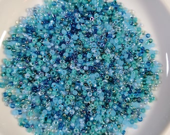 May include: A white ceramic bowl filled with a variety of small, round beads in shades of blue, teal, and clear. The beads are scattered, creating a textured surface. The beads are likely for jewelry making or crafts.