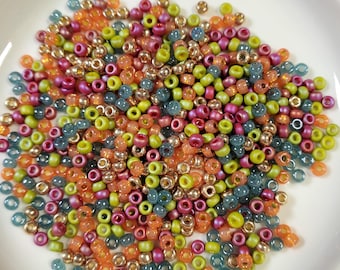May include: A collection of small, round glass beads in various shades of green, orange, pink, and gold. The beads are arranged in a pile on a white plate.