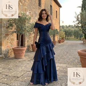 May include: A floor-length navy blue off-the-shoulder dress featuring a ruched bodice, a sweetheart neckline, and a multi-layered ruffled skirt. The garment is crafted from a shimmering fabric and includes a small black clutch held at the side.