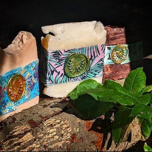 May include: Three bars of handmade soap, each with a unique appearance. One is light brown, the second is white, and the third is dark brown. Each bar is wrapped with a decorative band and a gold-coloured seal. Green leaves are in the foreground.