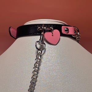 May include: Black and pink collar with a heart charm and a silver chain. The collar is made of black leather with pink accents and a pink heart-shaped charm. The chain is silver and attached to the collar with a clasp.
