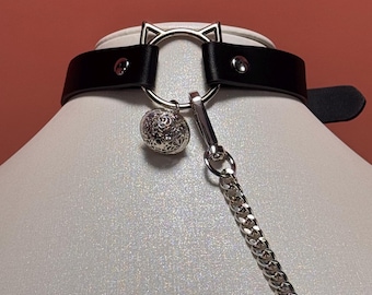 May include: Black leather choker with a silver cat ear design and a silver bell charm. The choker has a silver chain and a buckle closure. The background is a solid red-orange color.