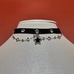 Puede incluir: Un collar tipo gargantilla negro con ojales metálicos plateados a lo largo de la banda. Una delicada cadena plateada cuelga debajo, adornada con pequeños eslabones en forma de corazón y un colgante central de estrella con centro negro.