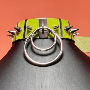 Puede incluir: Un collar de color verde lima con púas metálicas plateadas y detalles rectangulares verticales. El centro del collar muestra dos anillos metálicos plateados superpuestos unidos a una placa metálica vertical central.