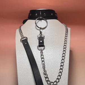 Puede incluir: Collar gargantilla negro con un gran anillo plateado y detalle de cadena. La gargantilla está hecha de cuero sintético negro y presenta remaches plateados. Una larga correa negra y una cadena plateada cuelgan del anillo.