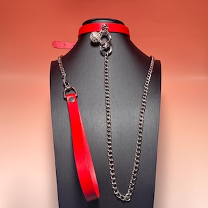 Puede incluir: Un collar rojo brillante con un anillo de metal plateado y un dije esférico colgante incrustado con cristales transparentes. Una larga cadena de metal plateado conecta el collar a una correa roja a juego con un cierre de metal plateado.