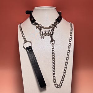 May include: Black leather choker necklace with silver chain and star charms. The necklace features a detachable leash with a large ring. The design includes multiple metal clasps and a decorative chain.