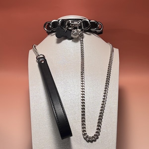 Puede incluir: Collar de cuero negro con herrajes de metal plateado, un dije en forma de hueso y un dije colgante. También se incluye una correa de cuero negro a juego con una cadena plateada. El collar y la correa se exhiben en una cabeza de maniquí blanca.