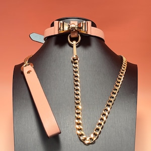 Puede incluir: Un collar de cuero rosa rubor con un lazo dorado y una correa de cadena dorada. El collar tiene una parte trasera negra y una lengüeta azul claro. La cadena dorada es gruesa y robusta.