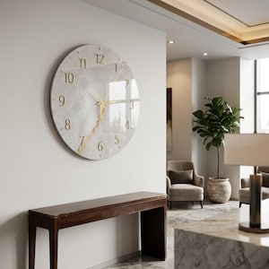 Puede incluir: Un gran reloj de pared circular con una esfera de efecto mármol gris claro, que presenta números dorados del 1 al 12 y manecillas a juego del mismo color. Una prominente veta dorada atraviesa diagonalmente el centro de la esfera.