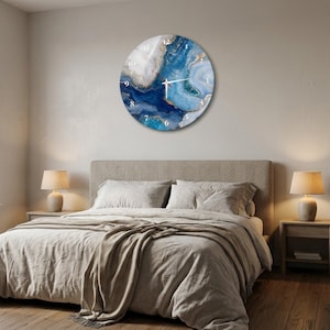 May include: A round wall clock featuring an abstract design with swirling shades of deep blue, light blue, and white, accented by gold-toned veins. The clock face displays white Arabic numerals and thin white hands, creating a circular timepiece.