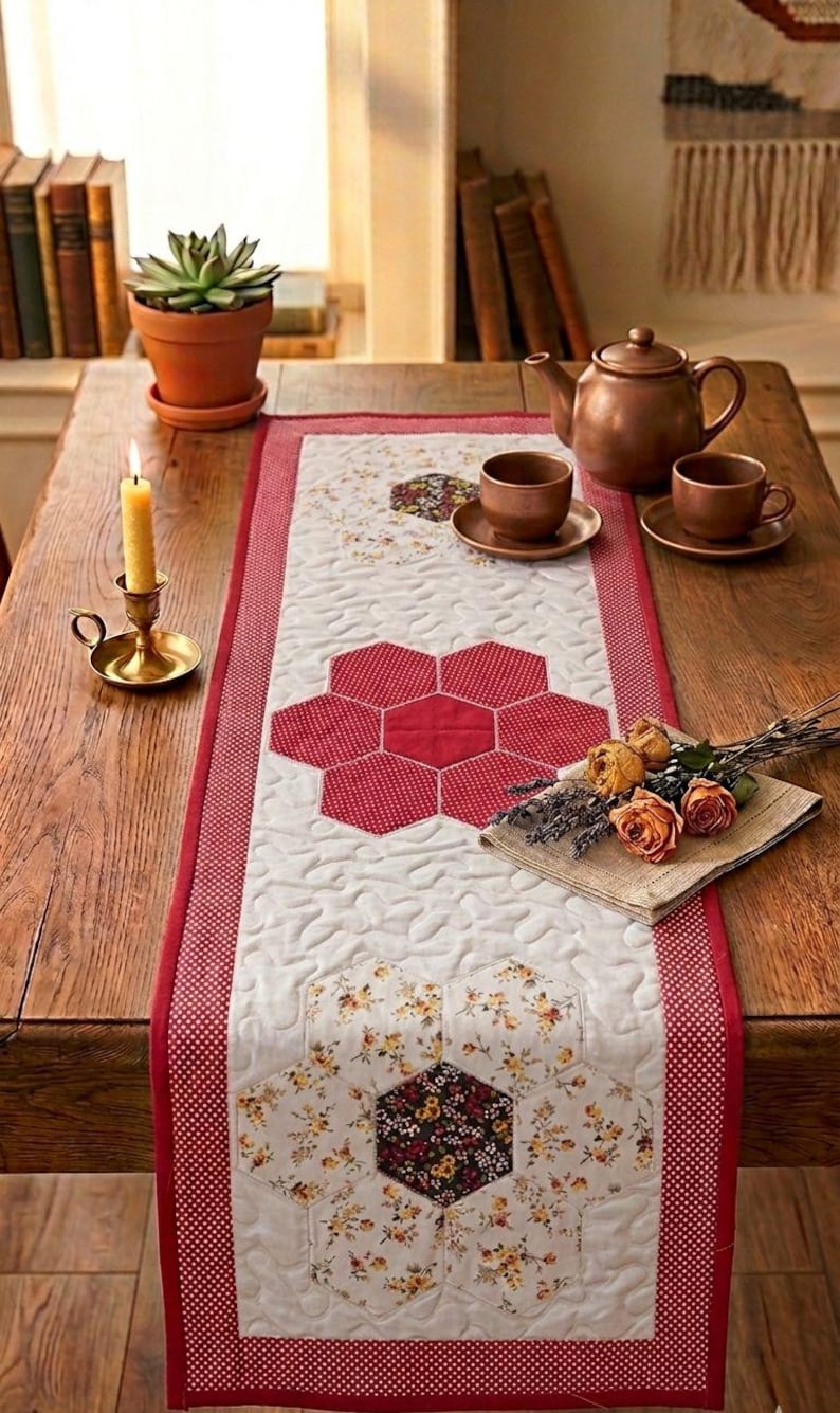 May include: A quilted table runner with a red and white floral design. The runner features a central pattern of red hexagon shapes and is bordered by a red polka dot fabric. A copper teapot, cups, and a candle are on the wooden table.