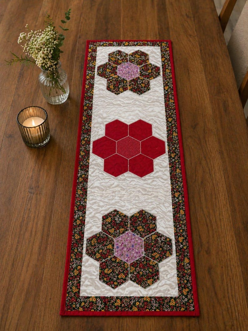May include: A quilted table runner with a white central panel and a red border. The runner features three large, flower-shaped designs. The flowers are made of red, floral, and purple fabric. The runner is placed on a wooden table.