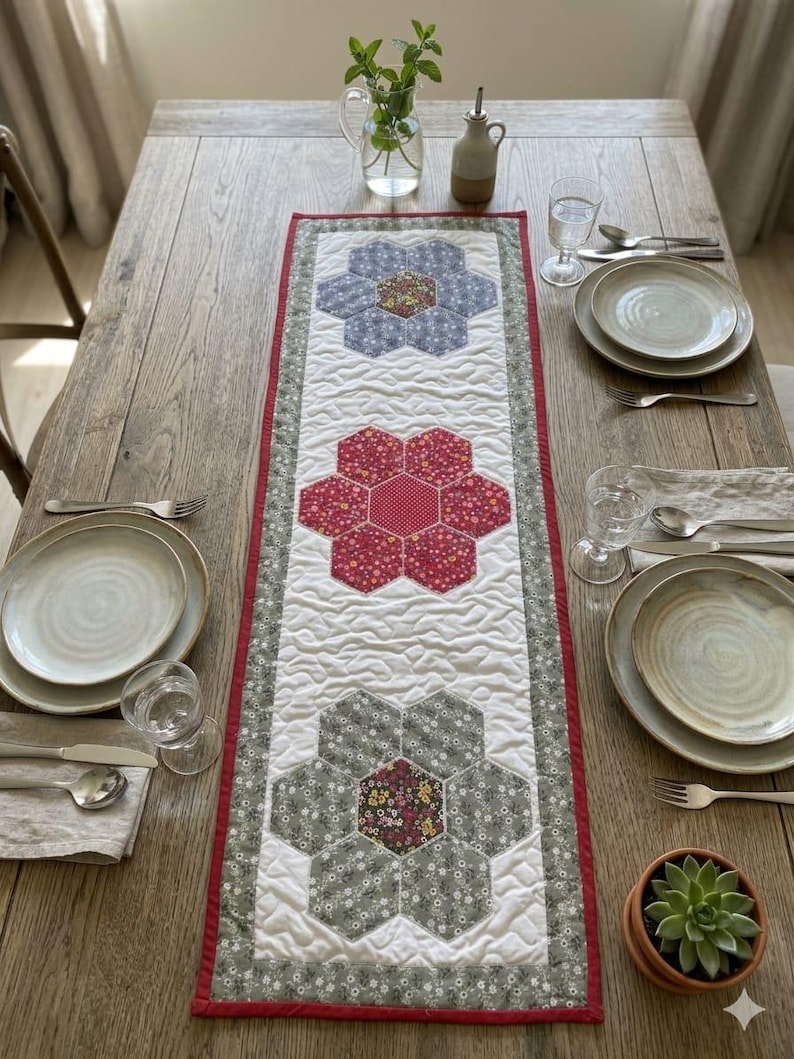 May include: A quilted table runner with three large floral designs in shades of blue, red, and gray. The runner has a white background with a red border and is placed on a wooden table. Table setting includes plates, silverware, and glasses.