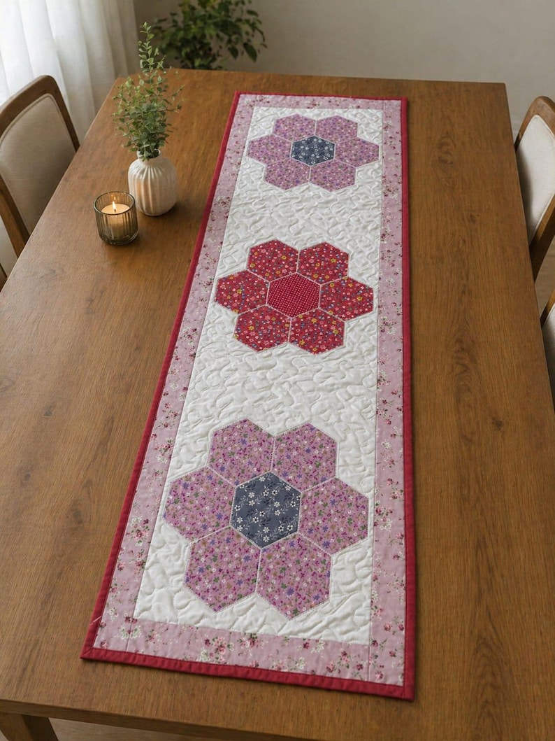 May include: A quilted table runner featuring three large flower designs. The flowers are made of various floral patterned fabrics in shades of pink, red, and blue, set against a white background. The runner has a red border and is placed on a wooden table.