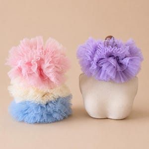 May include: A stack of three ruffled tulle collars in pink, cream, and blue. A separate lavender tulle collar is displayed on a mannequin head. The collars are made of soft, layered tulle fabric, creating a fluffy, textured appearance.