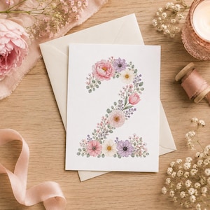 May include: A white rectangular greeting card featuring the number two illustrated with a variety of watercolor flowers including pink peonies, purple daisies, and small green leaves, resting on a light-colored wooden surface with pink ribbon.