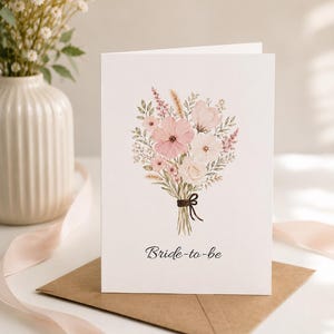 May include: A white card with a watercolor illustration of a floral bouquet in shades of pink and cream, tied with a brown ribbon. The words "Bride-to-be" are printed in black script below the bouquet. The card rests on a brown envelope.