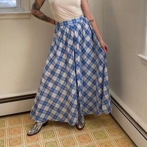 May include: A woman in a cream t-shirt and a long blue and white plaid skirt with a button-up front. She wears patterned boots. The room has a window and a patterned floor. The skirt's length is approximately 90 cm.